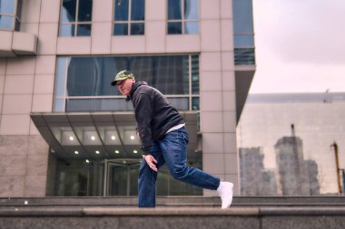 Formda bir genç adam coşkuyla break dans yapar, sert adam pozları verir, şınav çeker ve ritme ayak uydurur. Kot pantolon, sokak kıyafeti ve şapka giyiyor. Tavır, hareket ve enerji dolu. Sokak dansı, serbest stil ve modern dans..