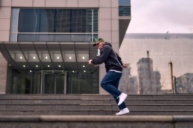 Formda bir genç adam coşkuyla break dans yapar, sert adam pozları verir, şınav çeker ve ritme ayak uydurur. Kot pantolon, sokak kıyafeti ve şapka giyiyor. Tavır, hareket ve enerji dolu. Sokak dansı, serbest stil ve modern dans..