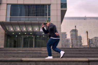 Formda bir genç adam coşkuyla break dans yapar, sert adam pozları verir, şınav çeker ve ritme ayak uydurur. Kot pantolon, sokak kıyafeti ve şapka giyiyor. Tavır, hareket ve enerji dolu. Sokak dansı, serbest stil ve modern dans..