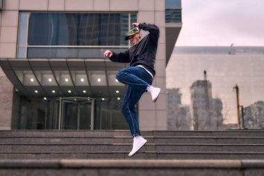A young adult dances hip hop breakdance, jumping and dancing enthusiastically in the streets. He wears comfortable clothes and exudes attitude, movement, and positive energy. Street dance, freestyle, modern dance.