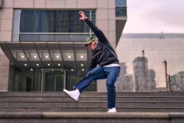 Formda bir genç adam coşkuyla break dans yapar, sert adam pozları verir, şınav çeker ve ritme ayak uydurur. Kot pantolon, sokak kıyafeti ve şapka giyiyor. Tavır, hareket ve enerji dolu. Sokak dansı, serbest stil ve modern dans..