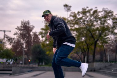 Genç, atletik bir adam şehirde coşkuyla dans ediyor, sert çocuk pozları veriyor ve ritme ayak uyduruyor. Kot pantolon, sokak kıyafetleri ve şapka giyiyor. Tavır, hareket ve iyi enerji yayıyor. Sokak dansı, serbest stil ve modern dans..