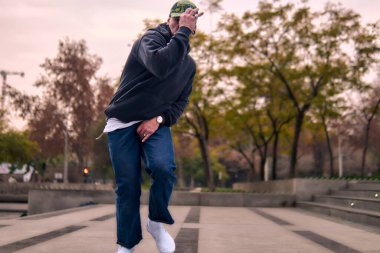 Genç, atletik bir adam şehirde coşkuyla dans ediyor, sert çocuk pozları veriyor ve ritme ayak uyduruyor. Kot pantolon, sokak kıyafetleri ve şapka giyiyor. Tavır, hareket ve iyi enerji yayıyor. Sokak dansı, serbest stil ve modern dans..