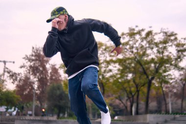 Genç, atletik bir adam şehirde coşkuyla dans ediyor, sert çocuk pozları veriyor ve ritme ayak uyduruyor. Kot pantolon, sokak kıyafetleri ve şapka giyiyor. Tavır, hareket ve iyi enerji yayıyor. Sokak dansı, serbest stil ve modern dans..