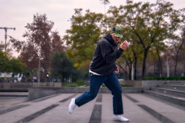 Genç, atletik bir adam şehirde coşkuyla dans ediyor, sert çocuk pozları veriyor ve ritme ayak uyduruyor. Kot pantolon, sokak kıyafetleri ve şapka giyiyor. Tavır, hareket ve iyi enerji yayıyor. Sokak dansı, serbest stil ve modern dans..
