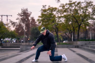 Genç, atletik bir adam şehirde coşkuyla dans ediyor, sert çocuk pozları veriyor ve ritme ayak uyduruyor. Kot pantolon, sokak kıyafetleri ve şapka giyiyor. Tavır, hareket ve iyi enerji yayıyor. Sokak dansı, serbest stil ve modern dans..