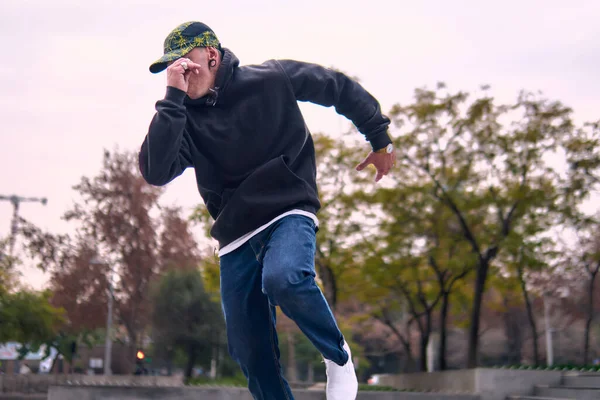 Genç, atletik bir adam şehirde coşkuyla dans ediyor, sert çocuk pozları veriyor ve ritme ayak uyduruyor. Kot pantolon, sokak kıyafetleri ve şapka giyiyor. Tavır, hareket ve iyi enerji yayıyor. Sokak dansı, serbest stil ve modern dans..
