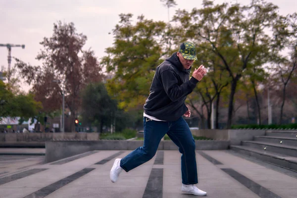 Genç, atletik bir adam şehirde coşkuyla dans ediyor, sert çocuk pozları veriyor ve ritme ayak uyduruyor. Kot pantolon, sokak kıyafetleri ve şapka giyiyor. Tavır, hareket ve iyi enerji yayıyor. Sokak dansı, serbest stil ve modern dans..