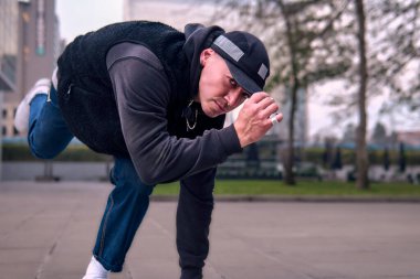 Yetişkin break dansçı hip hop, şehirde dans ve dönme, sokak kıyafetleri, kot pantolon ve şapka giyme, binalar arasında. Tarzı, duruşu ve enerjisi ile hareket ediyor..