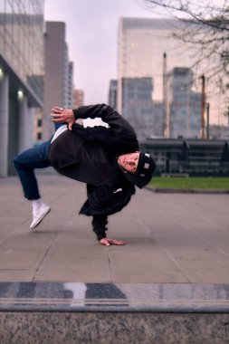 Yetişkin break dansçı hip hop, şehirde dans ve dönme, sokak kıyafetleri, kot pantolon ve şapka giyme, binalar arasında. Tarzı, duruşu ve enerjisi ile hareket ediyor..