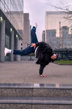 Yetişkin break dansçı hip hop, şehirde dans ve dönme, sokak kıyafetleri, kot pantolon ve şapka giyme, binalar arasında. Tarzı, duruşu ve enerjisi ile hareket ediyor..