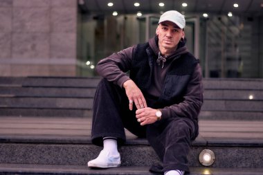 Yetişkin bir hip hop break dansçısının portresi, sakince poz veriyor, sokak kıyafeti, kot pantolon ve bir şapka giyiyor, bir binanın merdivenleri arasında oturuyor. Tarzı, duruşu ve enerjisi ile dans ediyor..