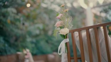 Açık havada yapılacak bir düğün için narin pembe çiçekler ve yeşillikle süslenmiş ahşap sandalyeye yakın çekim. Yumuşak doğal ışık ve bulanık bahçe arka planı.