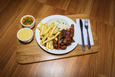 Peru yemeği Lomo Saltado, patates kızartması et, soğan, sarı biber, maydanoz, beyaz pirinç ve soya sosu, çatal bıçak takımı ve ahşap arka planda..