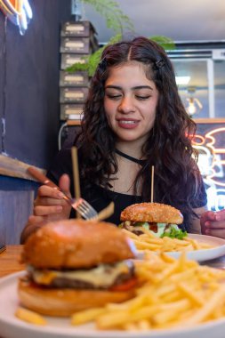 Genç kız restoranda oturup hamburger yiyor.