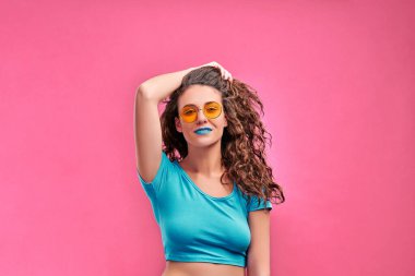 Uzun kıvırcık saçlı, moda güneş gözlüğü takan ve pembe arka planda izole bir şekilde kameraya bakan çekici kadının yakın görüntüsü. Boşluğu kopyala