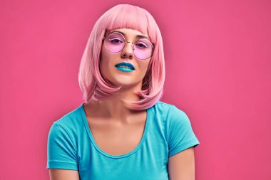 Pembe saçlı, moda güneş gözlüğü takan, pembe arka planda izole bir şekilde kameraya bakan çekici bir kadının yakın görüntüsü..