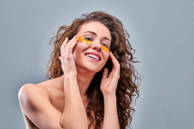 Genç ve güzel bir kız gözlerinin altına altın kolajen bantları yapıştırıyor. Kırışıklıkları ve koyu halkaları yok et. Kadın gözlerinin etrafındaki deriyle ilgilenir. Kozmetik prosedürleri. Yüz bakımı. İzole edilmiş gri arkaplan