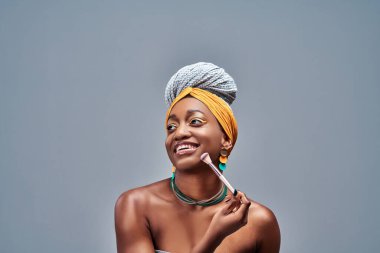 Pudralı fırça kullanan gülümseyen kız. Gri arka planda Afrikalı Amerikalı genç bir kızın fotoğrafı. Gençlik ve Güzellik
