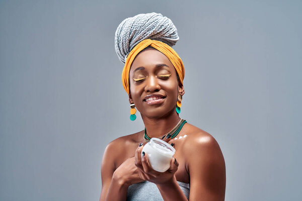 Beautiful young African American woman applying cream on skin on grey background. Copy space