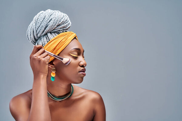 Young African woman in Studio doing body care and applying makeup, powder and rouge with a brush for young and beautiful skin. Side view. Copy space