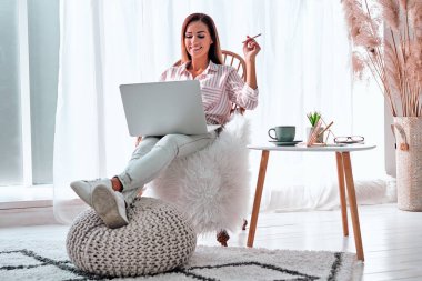 Evde iyi vakit geçiriyorum. Dizüstü bilgisayarda çalışan ve evinde rahat bir sandalyede otururken kahve içen genç ve güzel bir kadın. Düşük açılı çekim