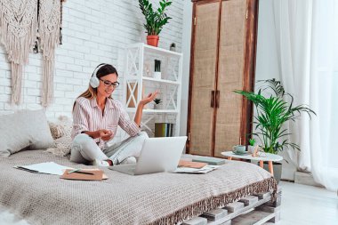 Çevrimiçi eğitim ve çalışma mesafesi. Kulaklıklı kadın video görüşmesi yapıyor. Mutlu ve gülümseyen bir kız evde, yatakta çalışıyor..