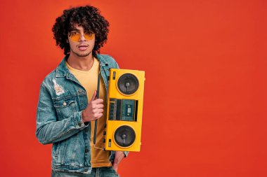 Sarı ritim kutusu olan şık gözlüklü genç bir Arap adam izole edilmiş arka planda nostaljik müzik dinliyor.