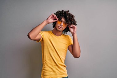 Gündelik kıyafetler giymiş genç seksi Arap adam şık sarı gözlüklerini ayarlıyor, ciddi bir şekilde kameraya bakıyor. Kendinden emin, zeki doğu öğrencisi.