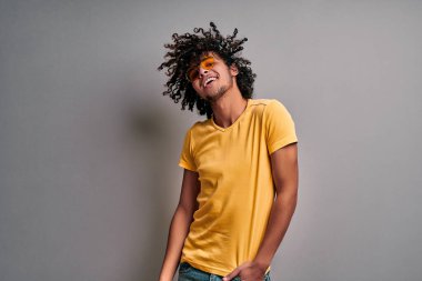 Gülümseyen, şık gözlüklü, neşeli, doğulu genç bir adam gri duvarda dalgalanan kıvırcık, egzotik saçlar. İzole edilmiş gri arka planda neşeli Arap öğrenci