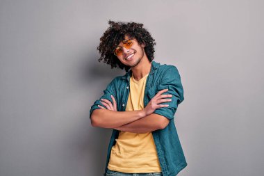 Gülümseyen mutlu doğu çocuğu ellerini çapraz yapıp gri duvara yaslanmış. Şık gözlüklü yakışıklı, neşeli Müslüman adam.