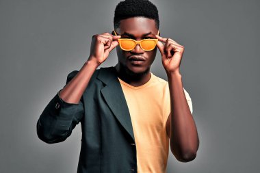 Yakışıklı genç adam gri arka planda dururken güneş gözlüğünü ayarlıyor. Tişört ve siyah ceket giyen kendine güvenen sakallı adam kameraya ciddi görünüyor..