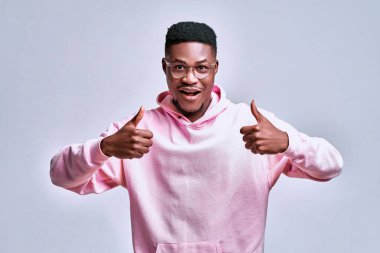 Yakışıklı bir Afro-Amerikalı küçük gözlüklerle gülümsüyor, pembe bir kapüşonlu giyiyor ve gri bir arka planda stüdyoda başparmağını gösteriyor..