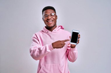Pembe kapüşonlu ve gözlüklü neşeli Afro-Amerikan adam bir telefon tutuyor ve ekranında gösteriyor. Cihazlar ve reklamlar kavramı.