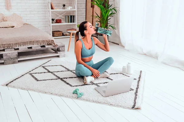 Young fit woman in sportswear drinks water while doing everyday morning warm up exercises on the floor at home, sportive lifestyle. Copy space