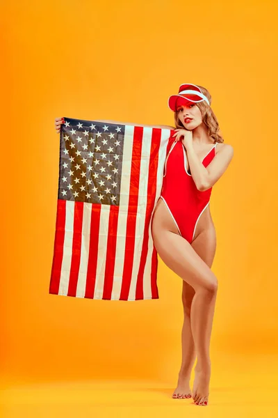 Vertical image of beautiful young woman in red underwear with flag United States of America on orange background. 4th july celebration concept