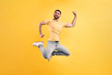 Zıplayarak dans edelim! Sarı tişört ve açık renk kot giyen genç adam güçlü elleri gösteren sarı bir duvarın arkasına atlıyor..