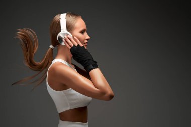 Young sport woman with headphones standing on grey background. Female boxer exercising on grey background. Side view. Copy space. clipart