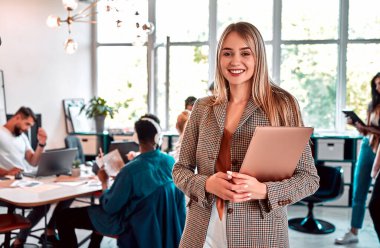 Ofis ortamında elinde dizüstü bilgisayarla oldukça neşeli bir iş kadınının portresini yakından çek. Geçmişi araştıran insanlar. Arkaplanda büyük panoramik pencere