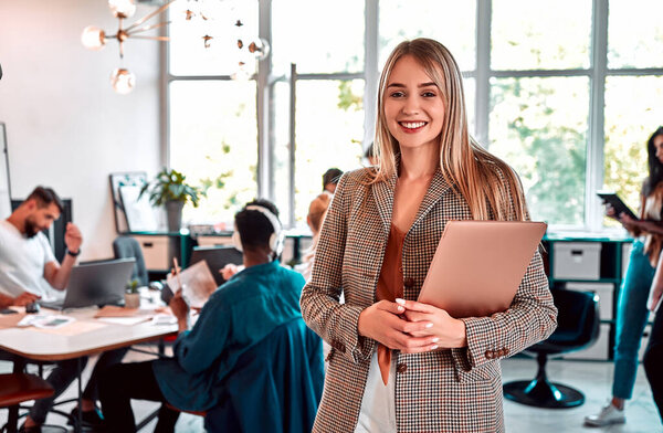 Close up view portrait of pretty cheerful business woman in an office environment holding laptop. People working background. Large panoramic window on background