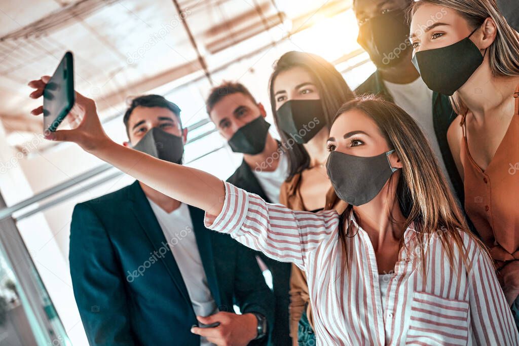 Young positive pleased business people colleagues indoors in office take a selfie wearing masks because of coronavirus. Side view. Selective focus