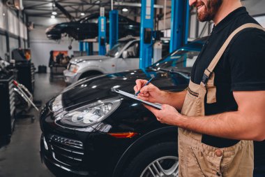 Üniformalı yakışıklı tamircinin otomobil servisinde dikilirken notlar alırken görüntüsü kesilmiş. Yan görünüm.