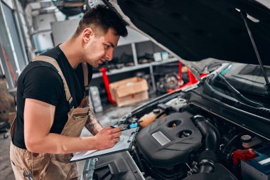 Araba servisi envanteri. Tamirhanedeki tamirci bir araba motorunu kontrol ederken panoya not bırakmış. Yan görünüm.