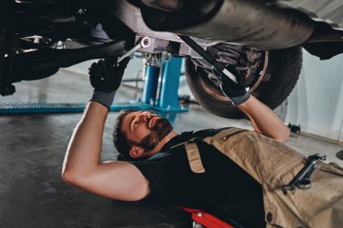 Üniformalı yakışıklı tamirci araba servisinde ingiliz anahtarlarıyla çalışıyor. Portatif plastik tamir sürüngeninde bir adam. Araba tamiri ve bakım konsepti.