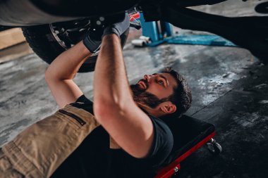 Yakışıklı bir erkek işçi araba sürüngeninin üzerinde yatıyor ve garajda otomobilin altında çalışırken LED kontrol lambası kullanıyor. Görüntüyü kapat.