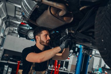 Araba tamircisi tamirhanedeki kaldırılmış otomobilin süspansiyonunu inceliyor. Görüntüyü kapat.