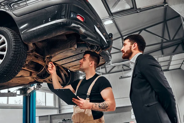 A professional mechanical employee of the car service workshop stand with the male customer under her car on the hydraulic ramp and show her defects and things they have to repair. Low angle view.