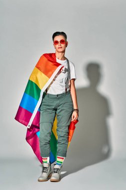 Happy beautiful woman in red eyewear smiling while posing with rainbow flag isolated over white background. Vertical photo.