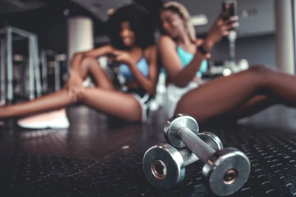 Shot Pair Metal Dumbbells Two African Girls Sitting Using Smartphone — Φωτογραφία Αρχείου