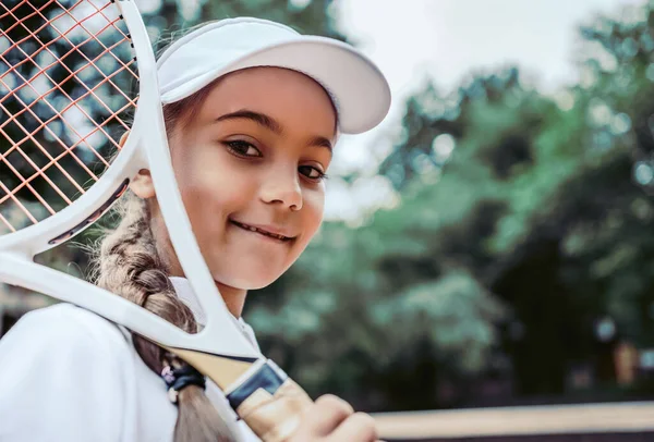 屋外で子供のためのテニストレーニング テニスコートで幸せなスポーティーな女の子の肖像画 白テニスの白人の子供はトレーニングでスポーツウェアを着用する スポーティ子供女の子見ますでザ カメラ — ストック写真